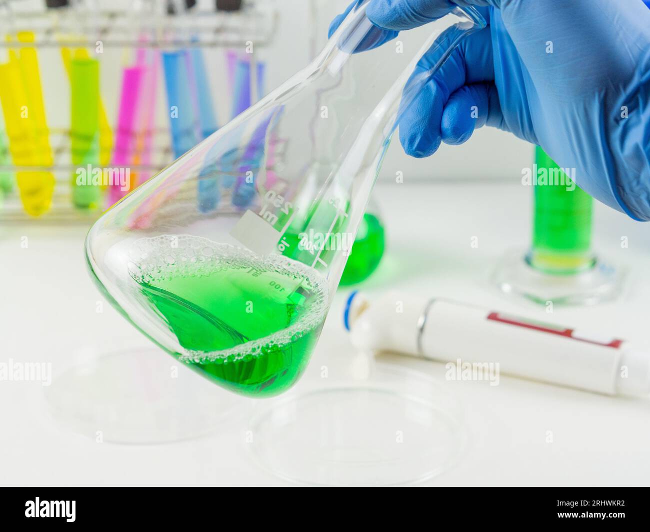 Nahaufnahme einer Hand mit Handschuhen, die einen erlenmeyerkolben mit einer grünen Flüssigkeitsprobe in einem Labor hält. Stockfoto