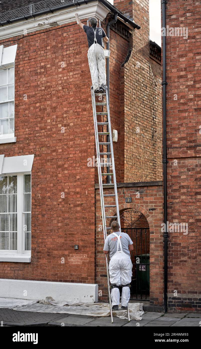 Hausmaler draußen arbeitet eine Leiter hinauf, um das Dach zu erreichen, während ein Kollege auf der Leiter steht, um zu verhindern, dass es verrutscht England UK Stockfoto