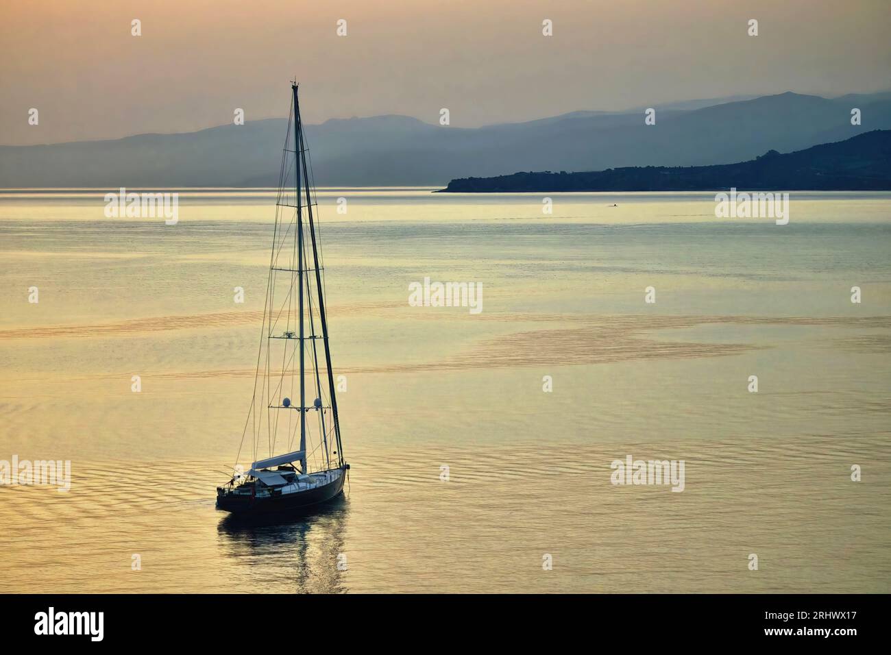 Eine Einzelyacht, die am frühen Morgen bei Sonnenaufgang auf ruhigem Wasser segelt, mit fernen Bergen und Hügeln in der Ferne Stockfoto