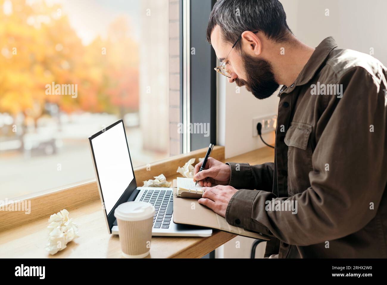 Bärtiger Geschäftsmann in lässigem Stil, der in einem Notizbuch in einem Café schreibt. Ein Mann sitzt in einem Café mit einem Laptop, Fernarbeit, Freiberufler, Herbst Stockfoto