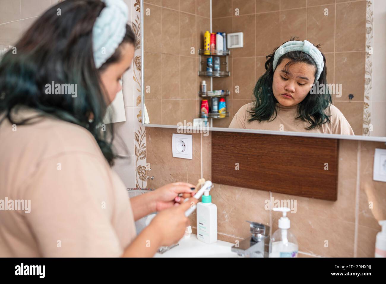 asiatisches Mädchen im Haarband, das die Zähne sauber macht Prothesen im Waschbecken vor dem Spiegel im Badezimmer, Routinekonzept am Morgen Stockfoto