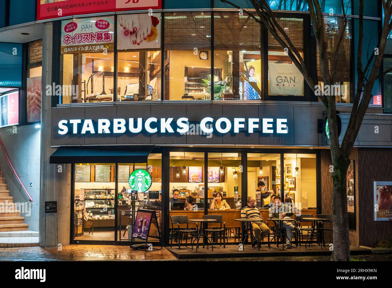 Das Starbucks Coffee Building in der Nacht befindet sich auf der ShiNGShopping Street in Kumamoto. Außenfassade aus Glas mit Personen, die innen und außen an Tischen sitzen Stockfoto