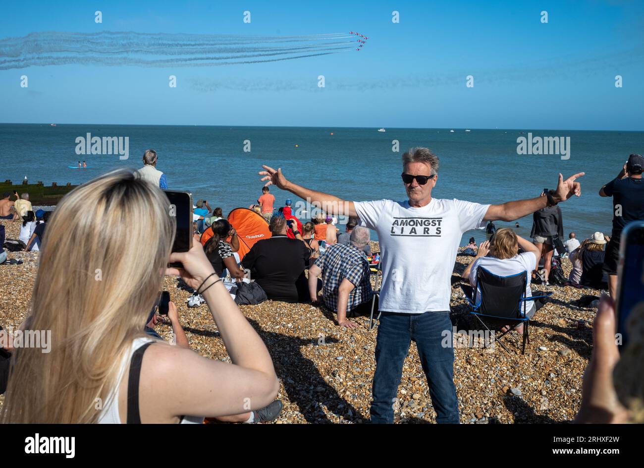 Eastbourne, Großbritannien. August 2023. Ein Mann, der ein T-Shirt mit der Aufschrift „Among Liars“ trägt, posiert für ein Foto am Eastbourne Beach, während das weltberühmte RAF-Showteam The Red Arrows auf der jährlichen Eastbourne Airbourne Airbourne, einer internationalen Flugshow, an der Küste vorbeifliegt. Die Show findet vier Tage lang statt, mit Flugvorführungen entlang der Küste. Stockfoto