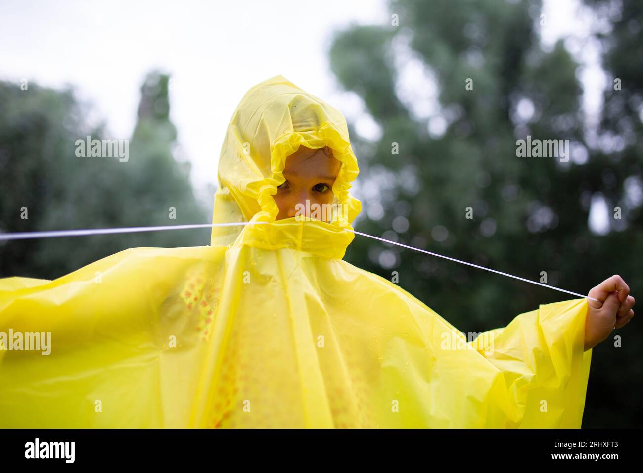 Nicht erkennbares Kind in gelbem Regenmantel, das bei regnerischem Wetter tagsüber vor unscharfen grünen Bäumen im Wald steht Stockfoto