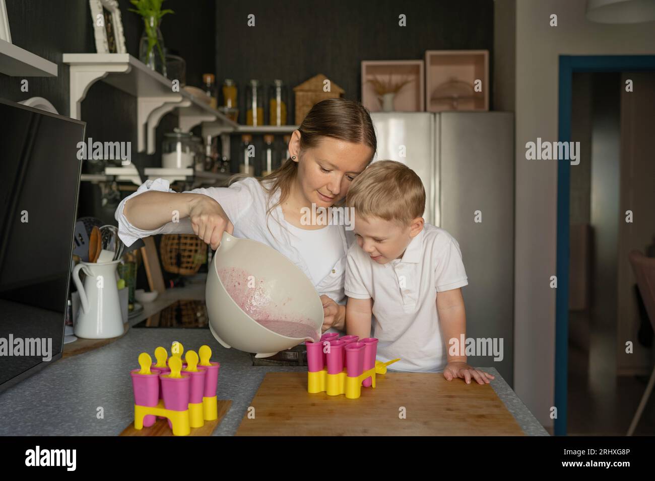 Kaukasischer Sohn mit blondem Haar, der neugierig auf die Mutter schaut, die Flüssigkeit in Eiswürfelformen gießt, während sie in der Küche in der Freizeit Eis zubereitet Stockfoto