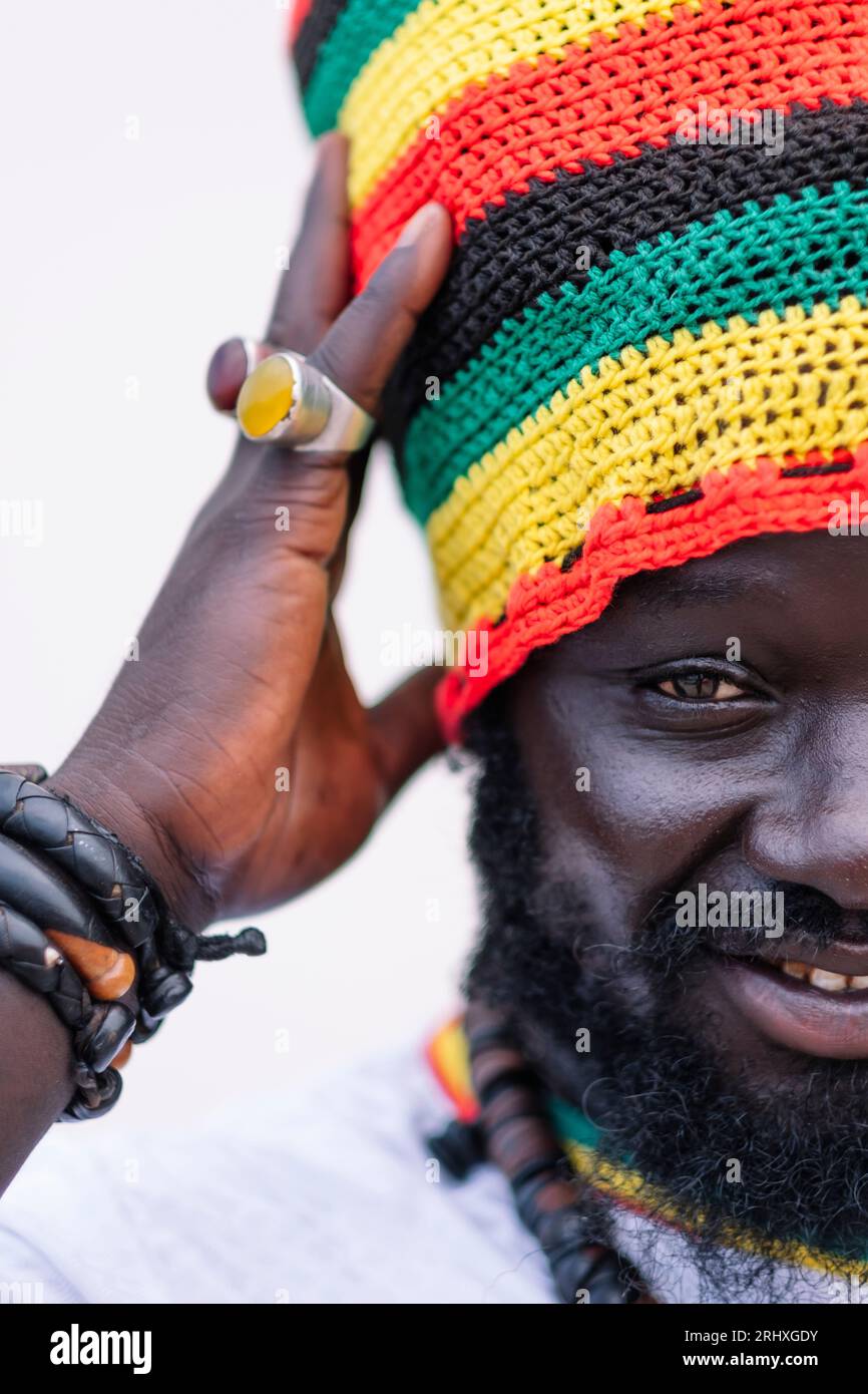 Porträt eines afroamerikanischen Mannes in einem bunten Hut, der steht und die Kamera anschaut, während er den Hut vor weißem Hintergrund berührt Stockfoto