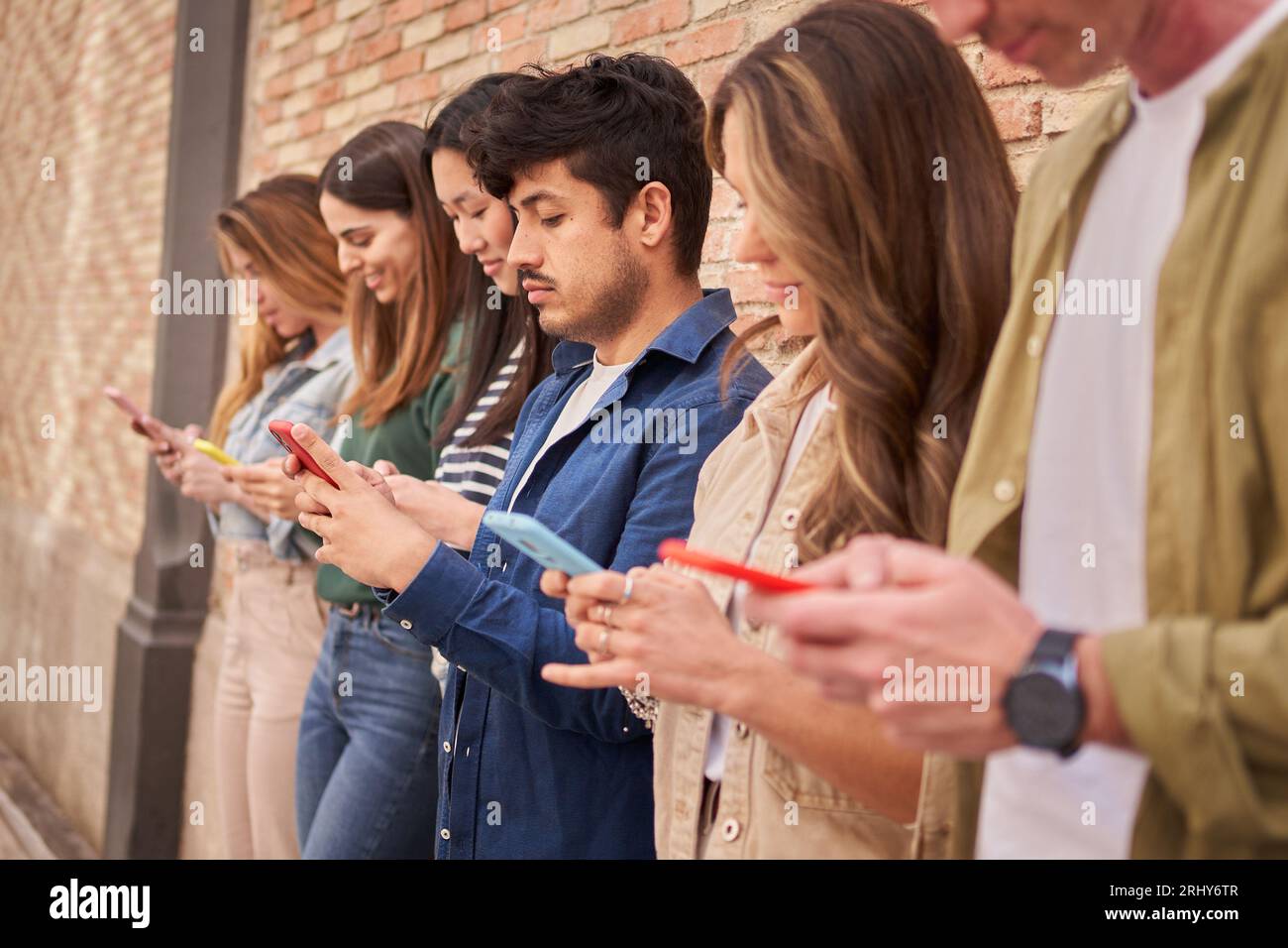 Seitenansicht multirassische Gruppe konzentrierte junge Generation z lehnte Ziegelwand mit Telefonen im Freien. Stockfoto