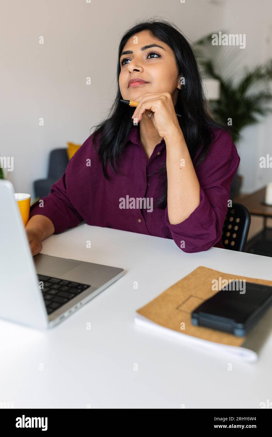 Nachdenkliche indische Frau, die mit Laptop auf dem Tisch sitzt, während sie wegschaut Stockfoto