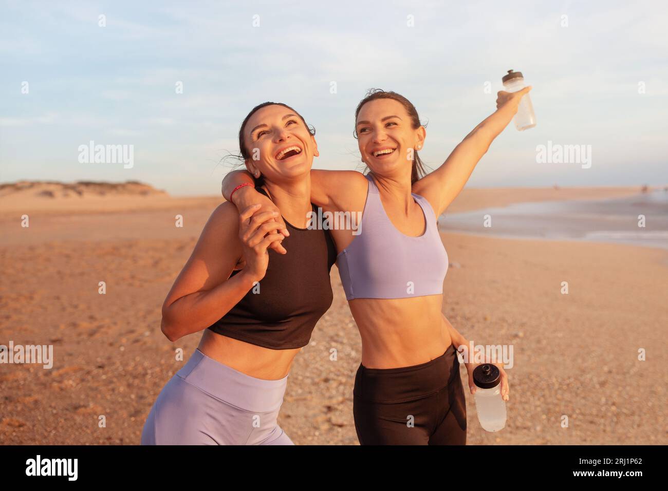 Zufriedene, fröhliche junge kaukasische Frauen umarmen sich, genießen morgens Workout, haben Spaß mit Wasserflaschen Stockfoto