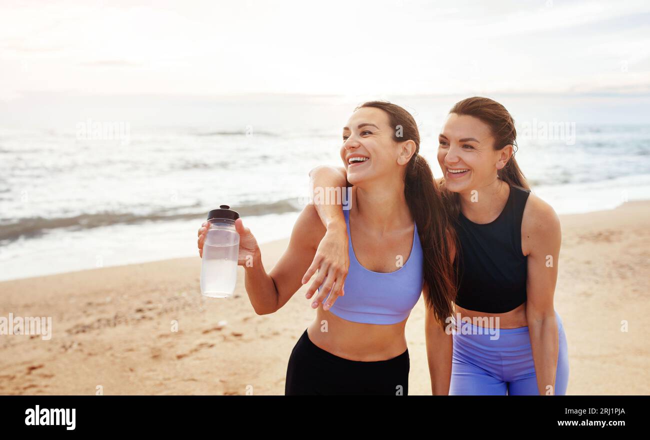Lachende, fröhliche junge kaukasierinnen, die sich mit Wasserflaschen umarmen, sich vom Workout erholen, viel Spaß haben Stockfoto