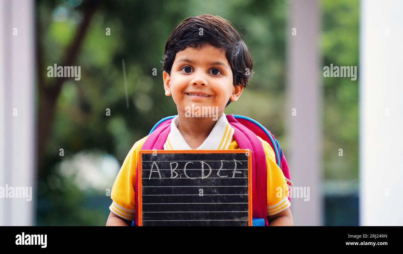 Portrait von glücklichen niedlichen kleinen indischen Jungen in der Schuluniform mit leerem Schiefer vor orangefarbenem Hintergrund, entzückende Grundschule Kind zeigt schwarze Tafel. Stockfoto