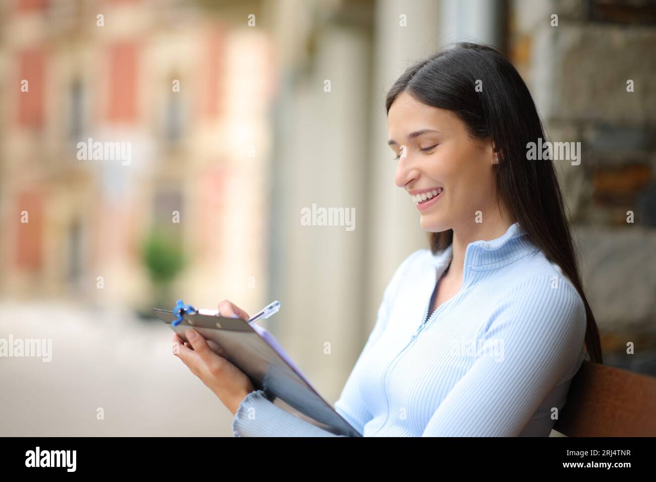 Glückliche Frau, die die Umfrage ausfüllt, sitzt auf einer Bank auf der Straße Stockfoto