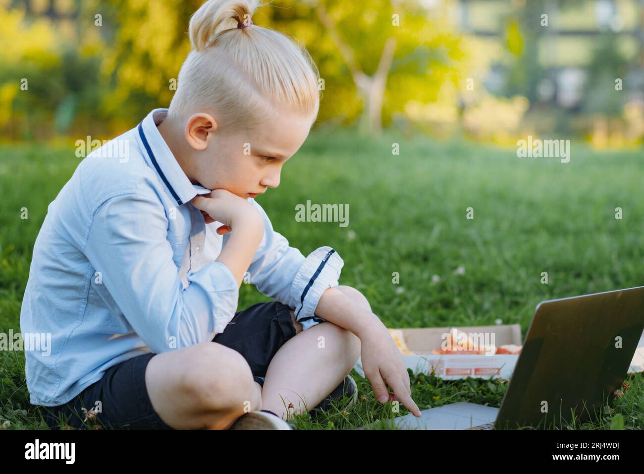 Niedlicher kaukasischer Junge mit Pferdeschwanz in blauem Hemd und Shorts, der im Park mit Laptop auf Gras sitzt. Hausaufgaben im Freien nach der Schule Stockfoto