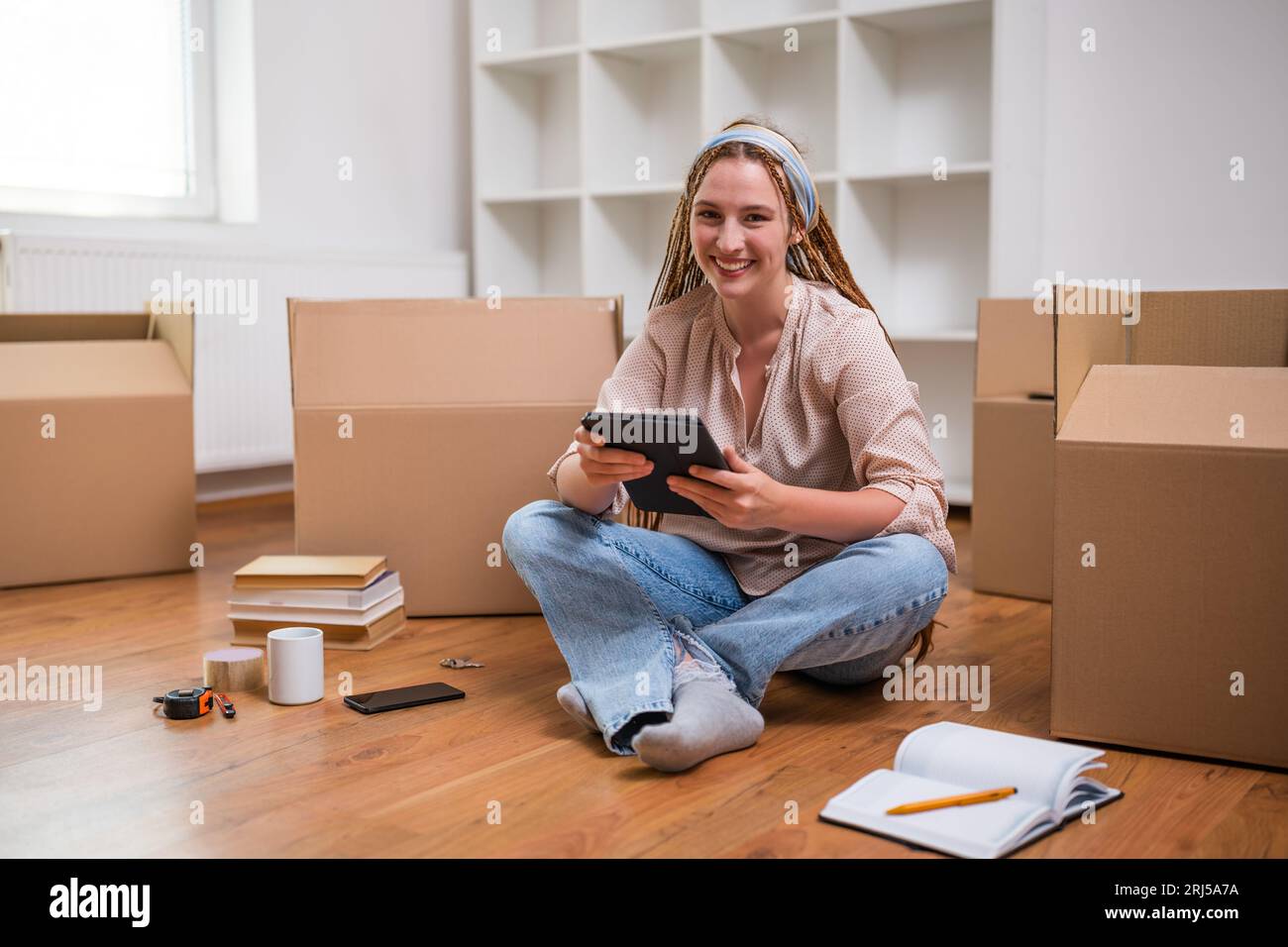 Moderne Ingwerfrau mit Zöpfen auf digitalem Tablet beim Einzug in eine neue Wohnung. Stockfoto