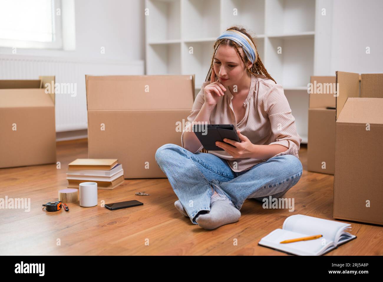 Moderne Ingwerfrau mit Zöpfen auf digitalem Tablet beim Einzug in eine neue Wohnung. Stockfoto