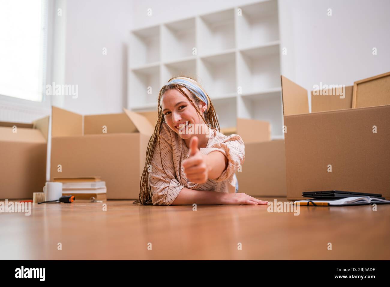 Moderne Ingwerfrau mit Zöpfen zieht in neue Wohnung und zeigt Daumen auf. Stockfoto