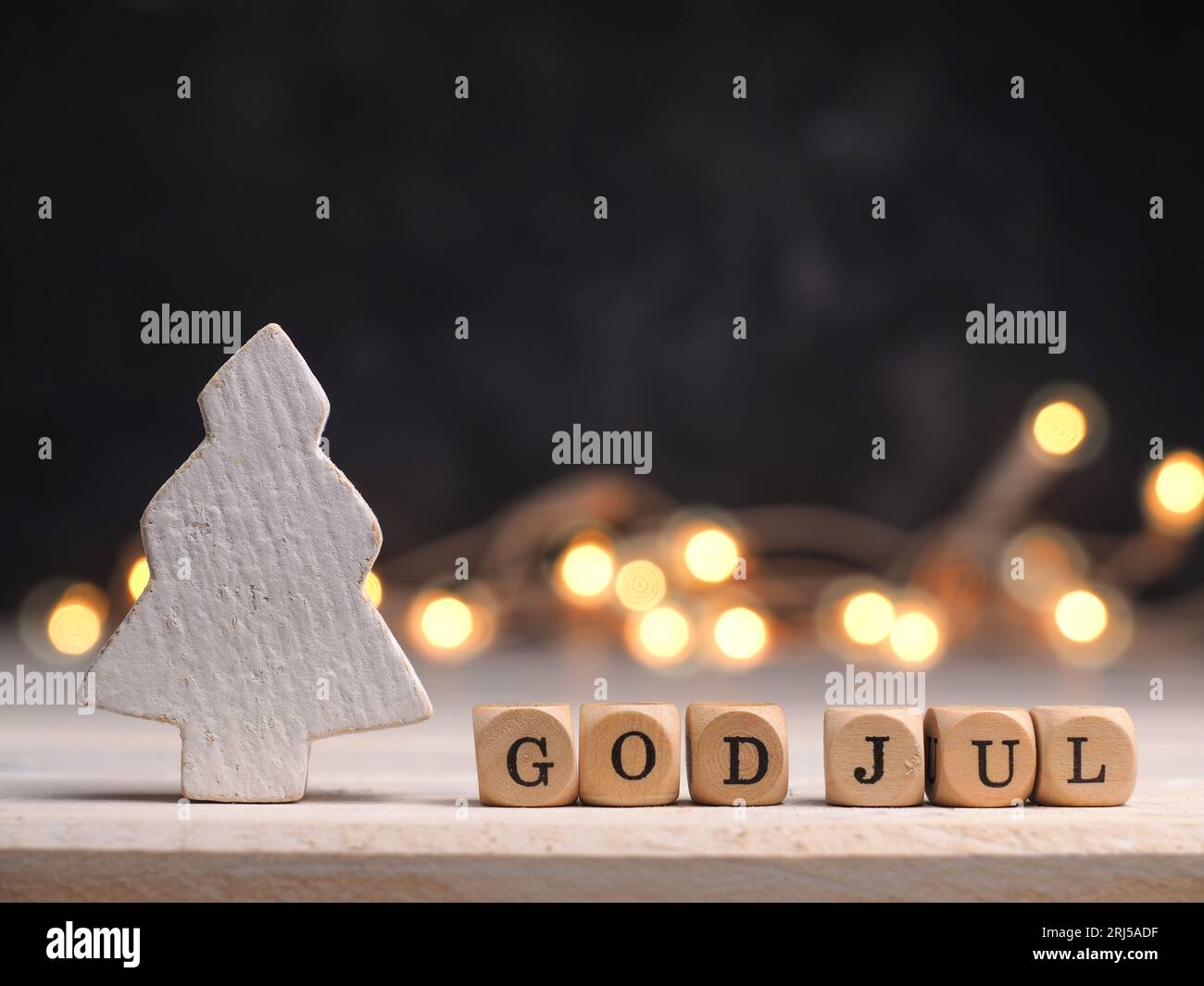 Kleine Holzblöcke mit God Jul und weißem hölzernem Weihnachtsbaum, verschwommene Lichter im Hintergrund Stockfoto
