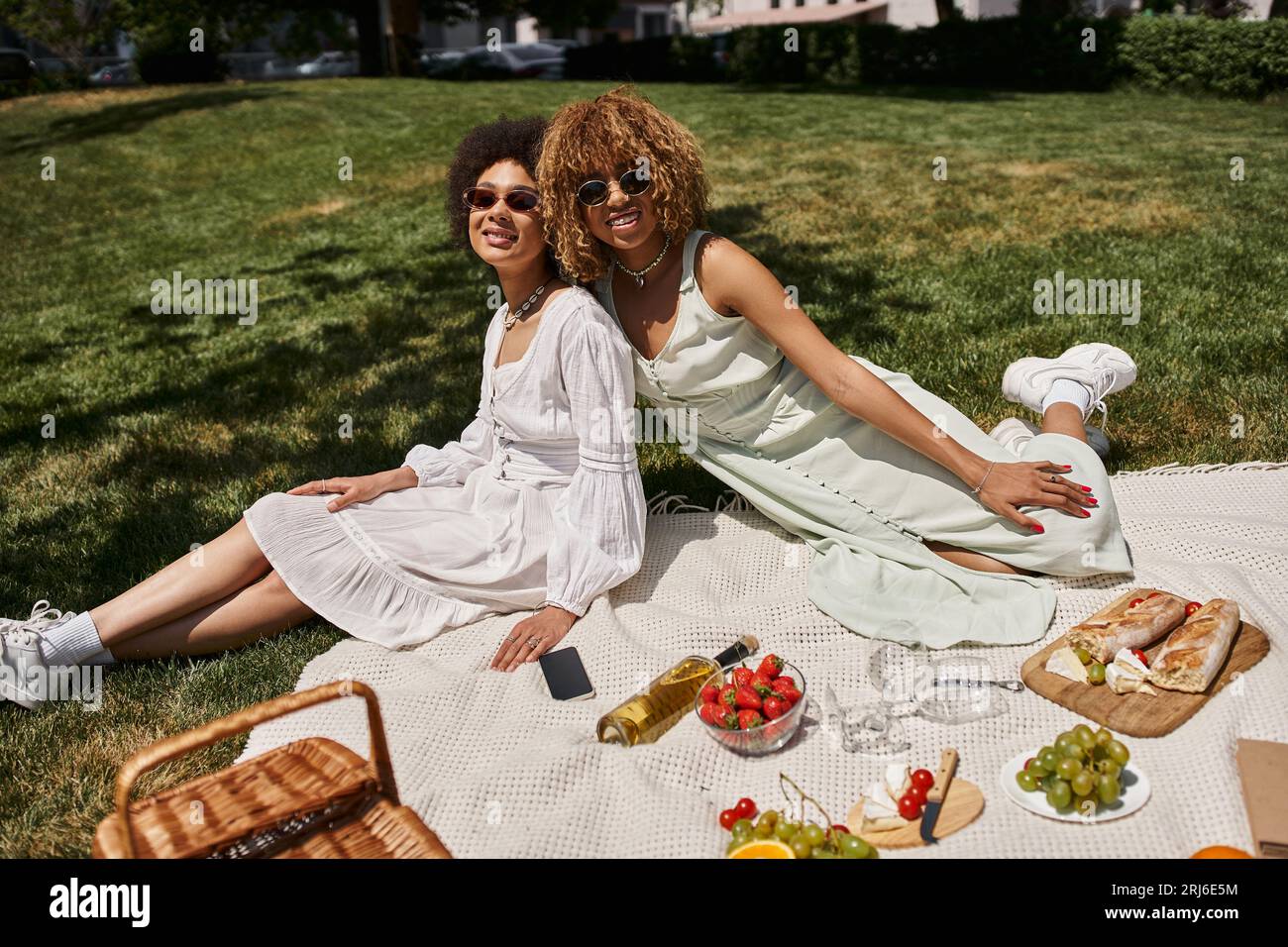 Stilvolle afroamerikanische Freundinnen, die sich auf einer Decke in der Nähe von Essen und Wein auf einem Sommerpicknick ausruhen Stockfoto