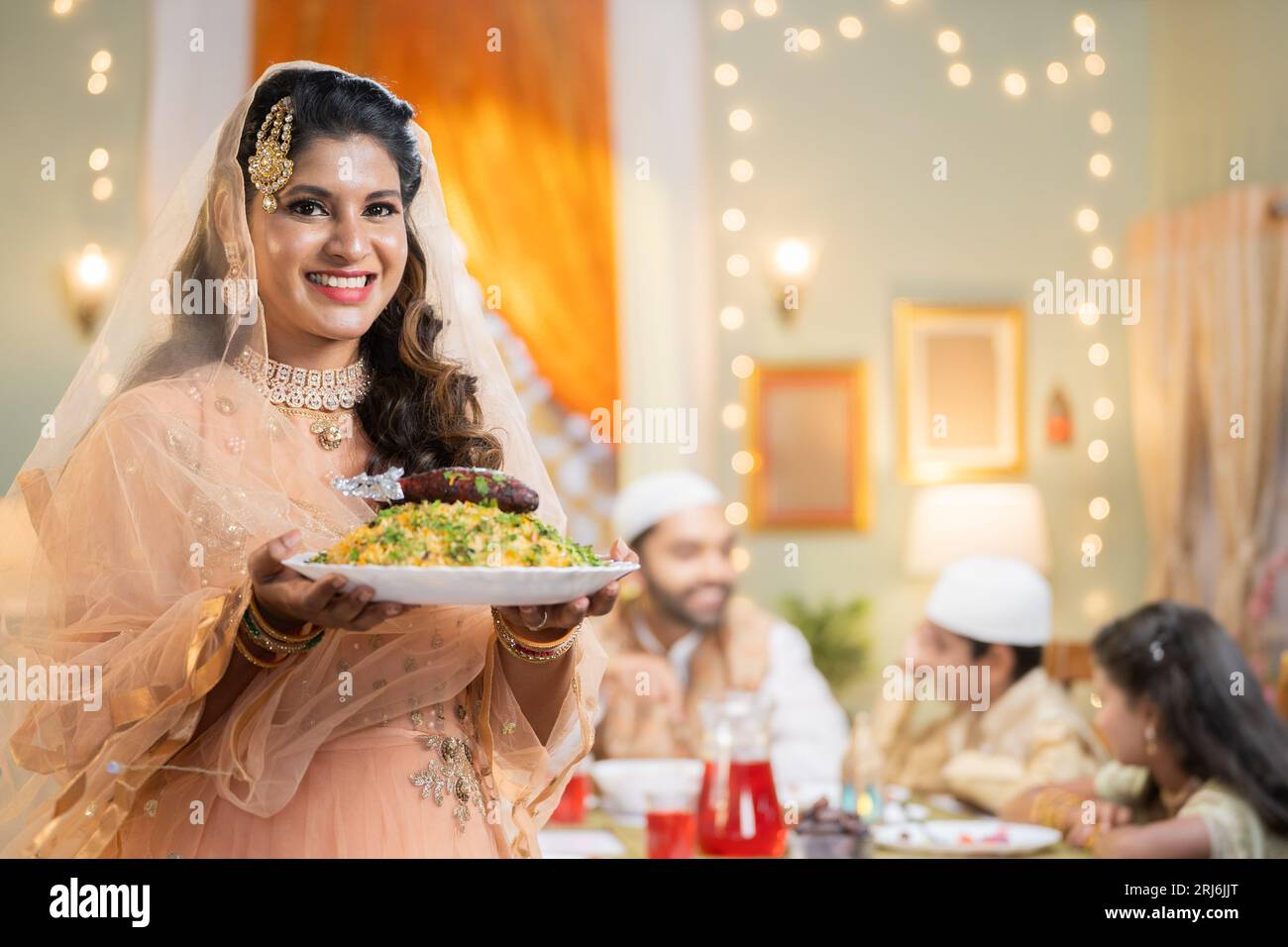 Glückliche lächelnde indische muslimische Frau, die Biryani-Teller in der Hand hält, indem sie die Kamera vor dem Ehemann mit Kindern anschaut, die sich zu Hause unterhalten - Konzept von Stockfoto