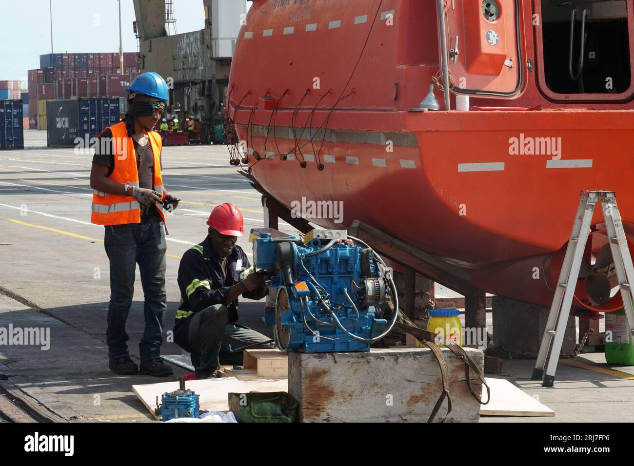 Zwei schwarze Mechaniker oder Techniker reparieren den Motor des orangefarbenen Rettungsbootes, der an der Ladestation befestigt ist. Stockfoto