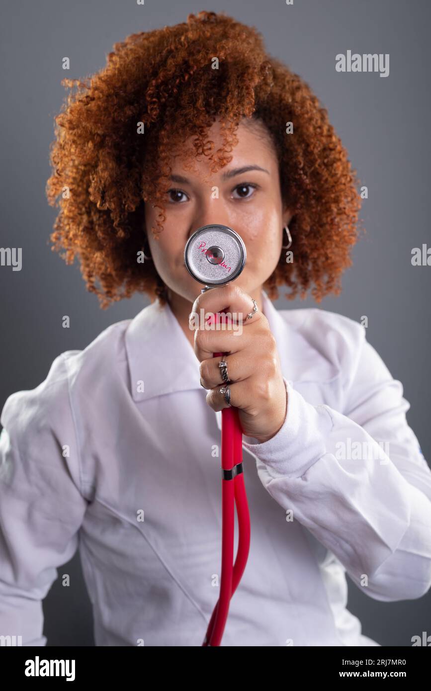 Porträt einer Tierärztin mit rotem Haar, die eine weiße Uniform trägt und ein Stethoskop nahe am Gesicht hält. Tierpflege. Isoliert auf grauem BA Stockfoto