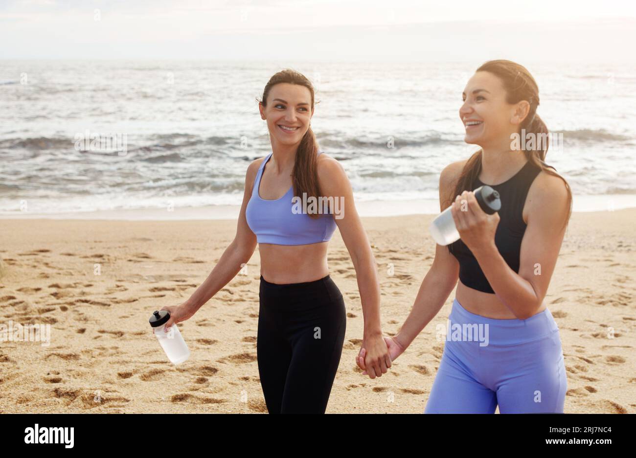 Glückliche junge kaukasische Frauen halten Hände, mit Flaschen Wasser, ruhen sich aus dem Workout aus, haben Spaß am Strand Stockfoto