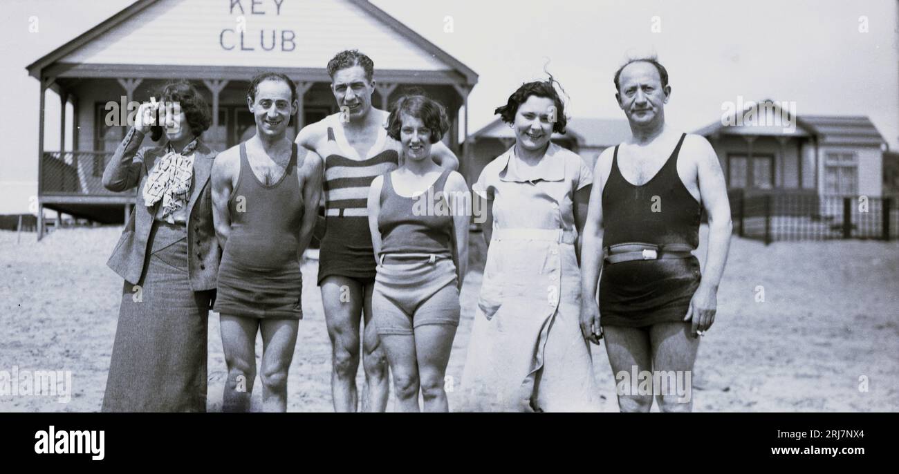 1920er Jahre, historisch, Sommer und eine Gruppe von Erwachsenen, die zusammen stehen, um ein Foto am Strand vor einem Holzchalet oder einer Hütte, Key Club, USA, zu machen. Das Bild zeigt die Bademode für Männer und Frauen aus dieser Zeit. Stockfoto