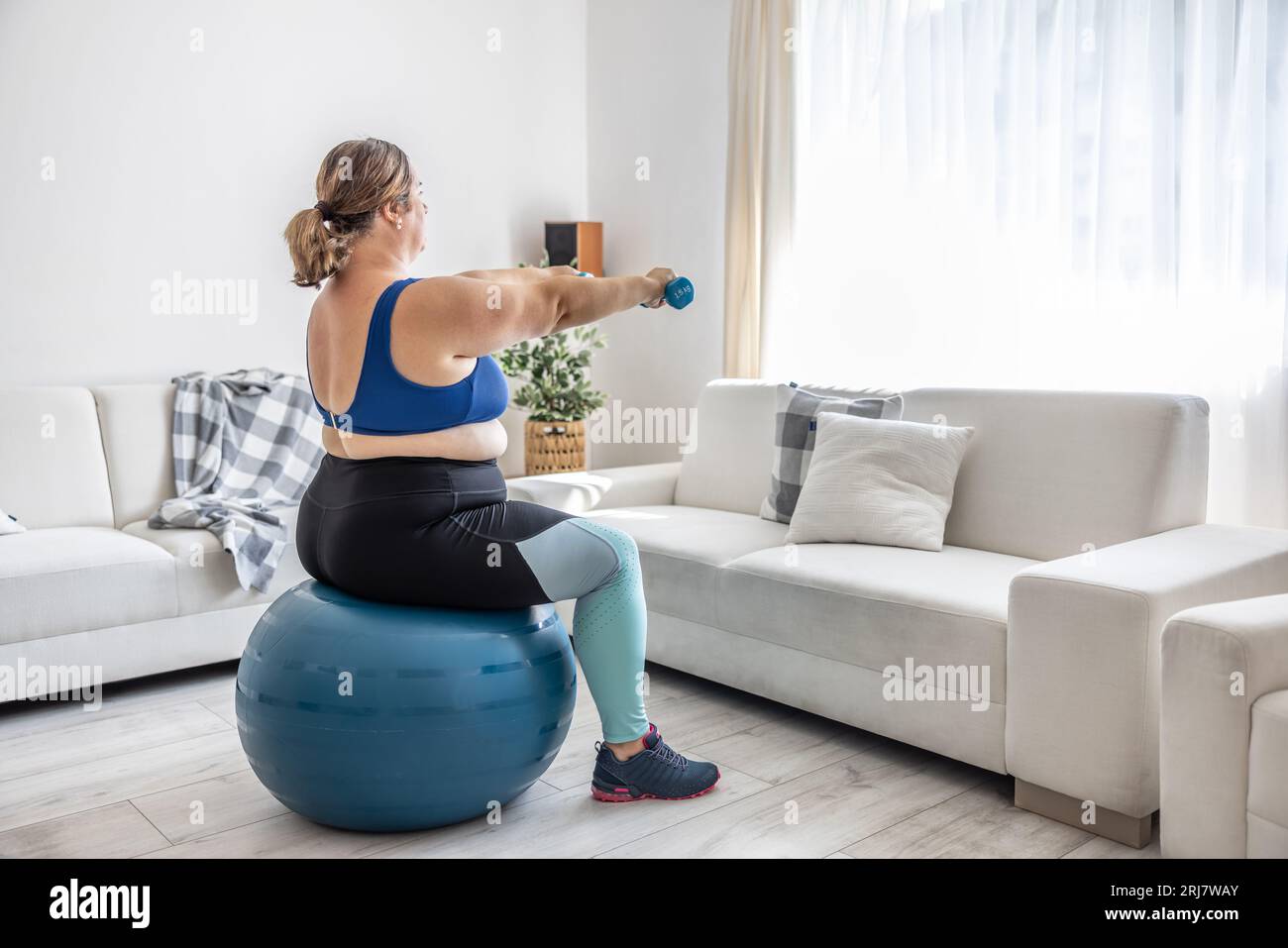 Übergewichtige Dame, die im Wohnzimmer Sport treibt. Er sitzt auf einem Fitnessball und hält seine Hände mit Kurzhanteln vor sich. Stockfoto