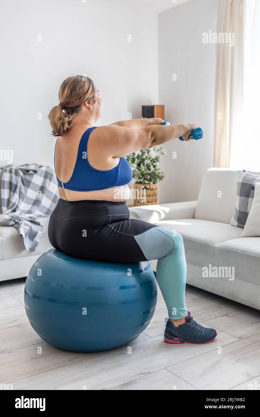 Übergewichtige Dame, die im Wohnzimmer Sport treibt. Er sitzt auf einem Fitnessball und hält seine Hände mit Kurzhanteln vor sich. Stockfoto