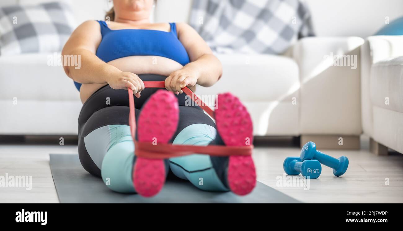 Übergewichtige Dame, die im Wohnzimmer Sport treibt. Er liegt auf der Matte und dehnt die Sportgummis. Stockfoto