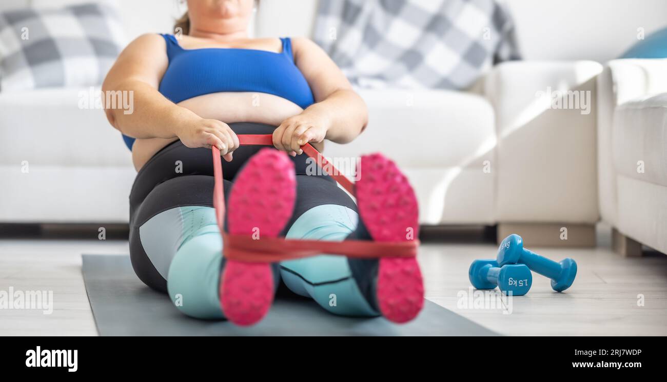 Übergewichtige Dame, die im Wohnzimmer Sport treibt. Er liegt auf der Matte und dehnt die Sportgummis. Stockfoto