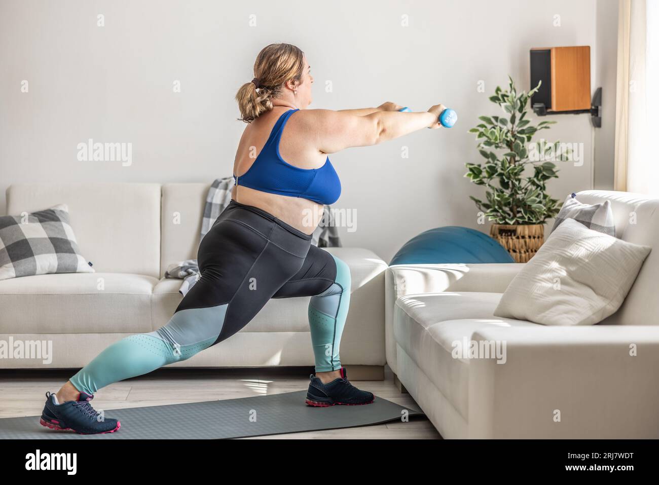 Übergewichtige Dame, die im Wohnzimmer Sport treibt. Er springt und hält Kurzhanteln in der Hand. Stockfoto
