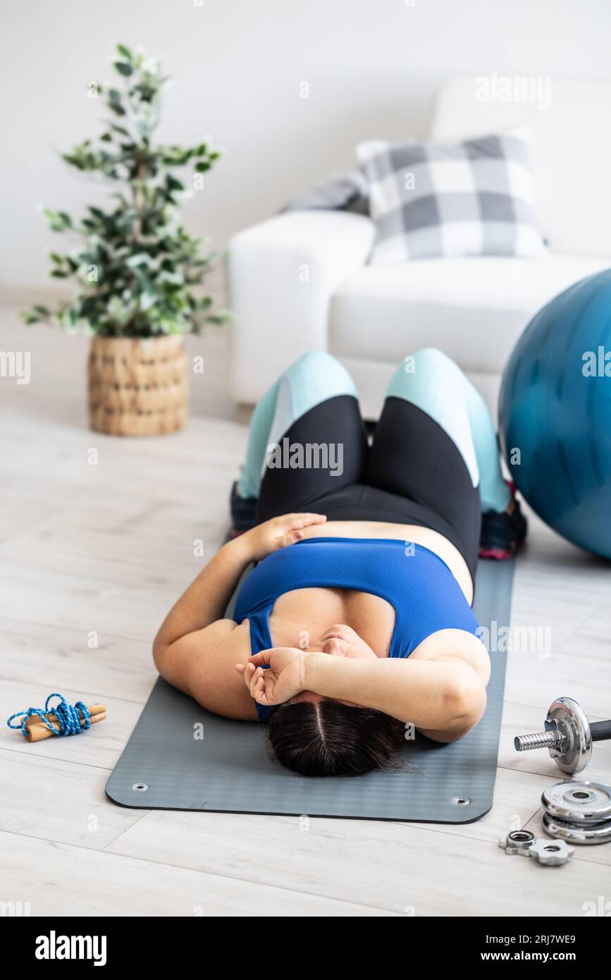 Eine müde Frau in Übergröße liegt auf einer Matte im Wohnzimmer, Sportausrüstung um sie herum. Stockfoto