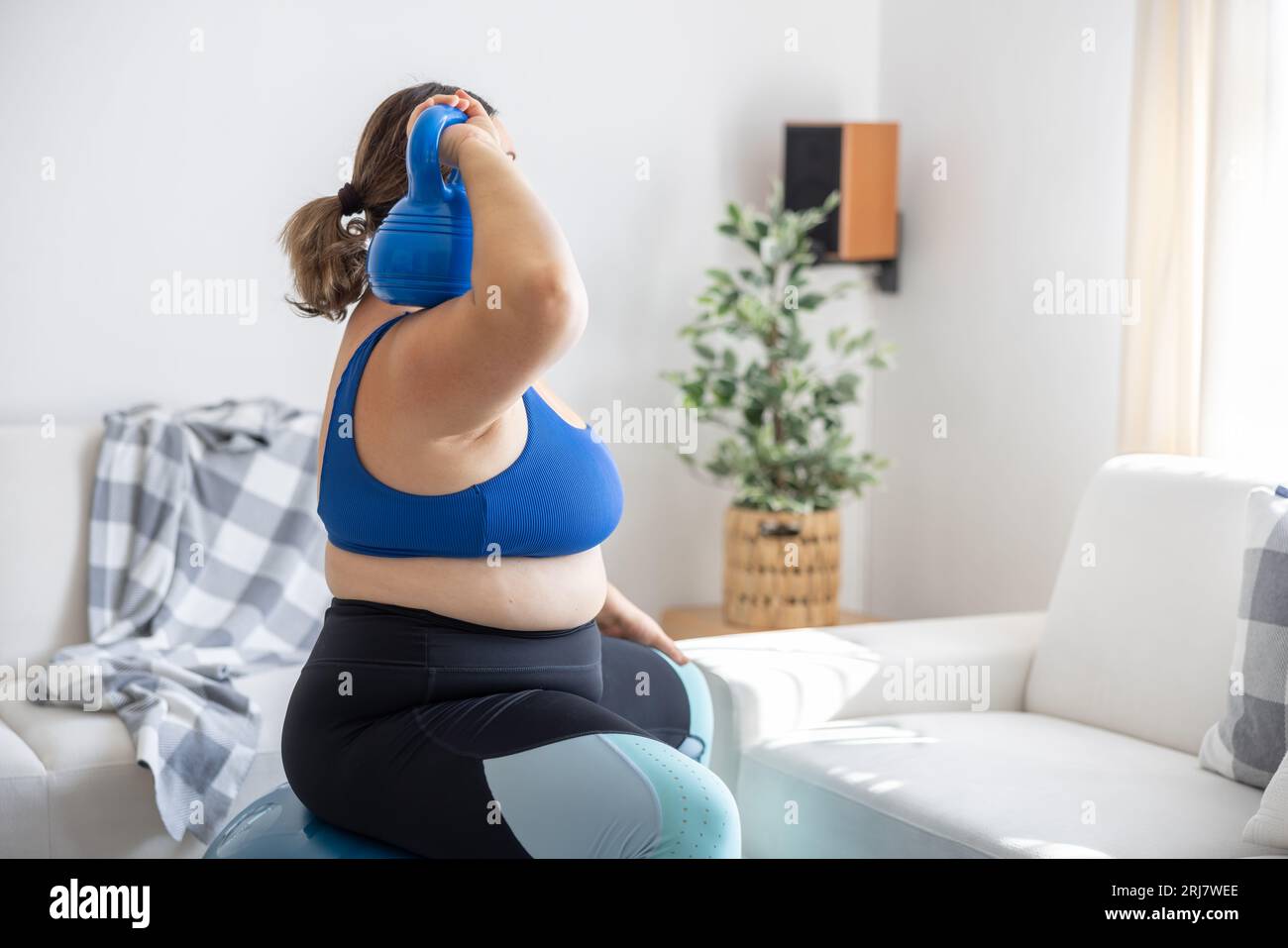 Übergewichtige Dame, die im Wohnzimmer Sport treibt. Sie sitzt auf einem Fitnessball und hält eine Kurzhantel auf der Schulter. Stockfoto
