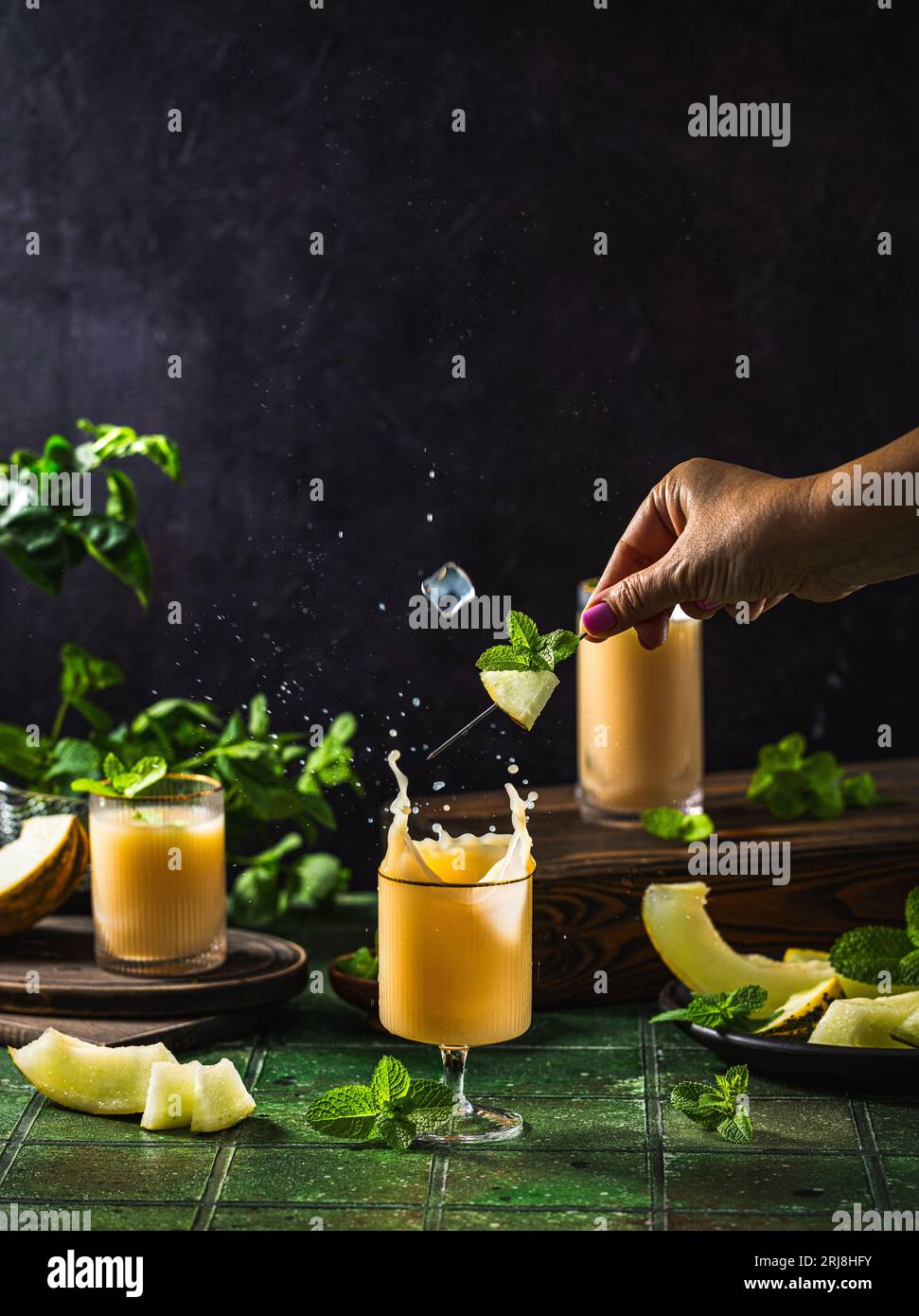 Frau bereitet Melonencocktail mit Minze im Glas auf dunkelgrünem Keramikfliesenhintergrund zu, Nahaufnahme. Getränke im Herbst und alkoholische Cocktails im Sommer. Alcoh Stockfoto