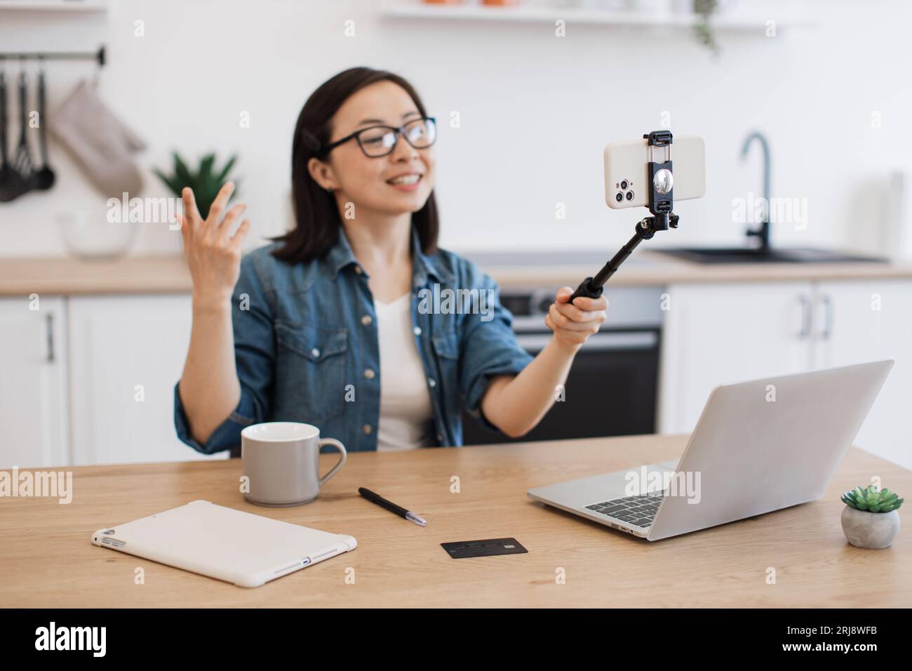 Eine energiegeladene Frau, die sich auf einem Selfie-Stick mit einem modernen Smartphone für einen persönlichen Blog filmt. Positive asiatische Frau in Brillen, die Online-Kurse aufnehmen wh Stockfoto