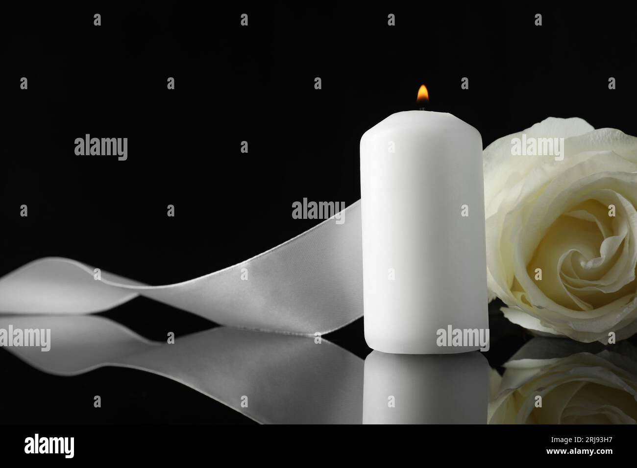 Brennende Kerze, weiße Rose und Band auf schwarzer Spiegeloberfläche in Dunkelheit, Nahaufnahme mit Platz für Text. Beerdigungssymbole Stockfoto