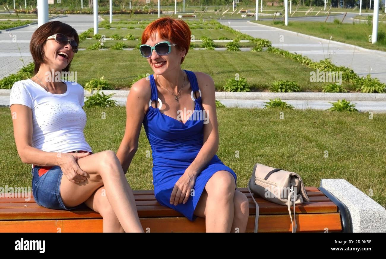 Zwei junge russische Schwestern in Sonnenbrille sitzen auf der Bank und lachen aufrichtig an einem Sommertag zusammen. Stockfoto