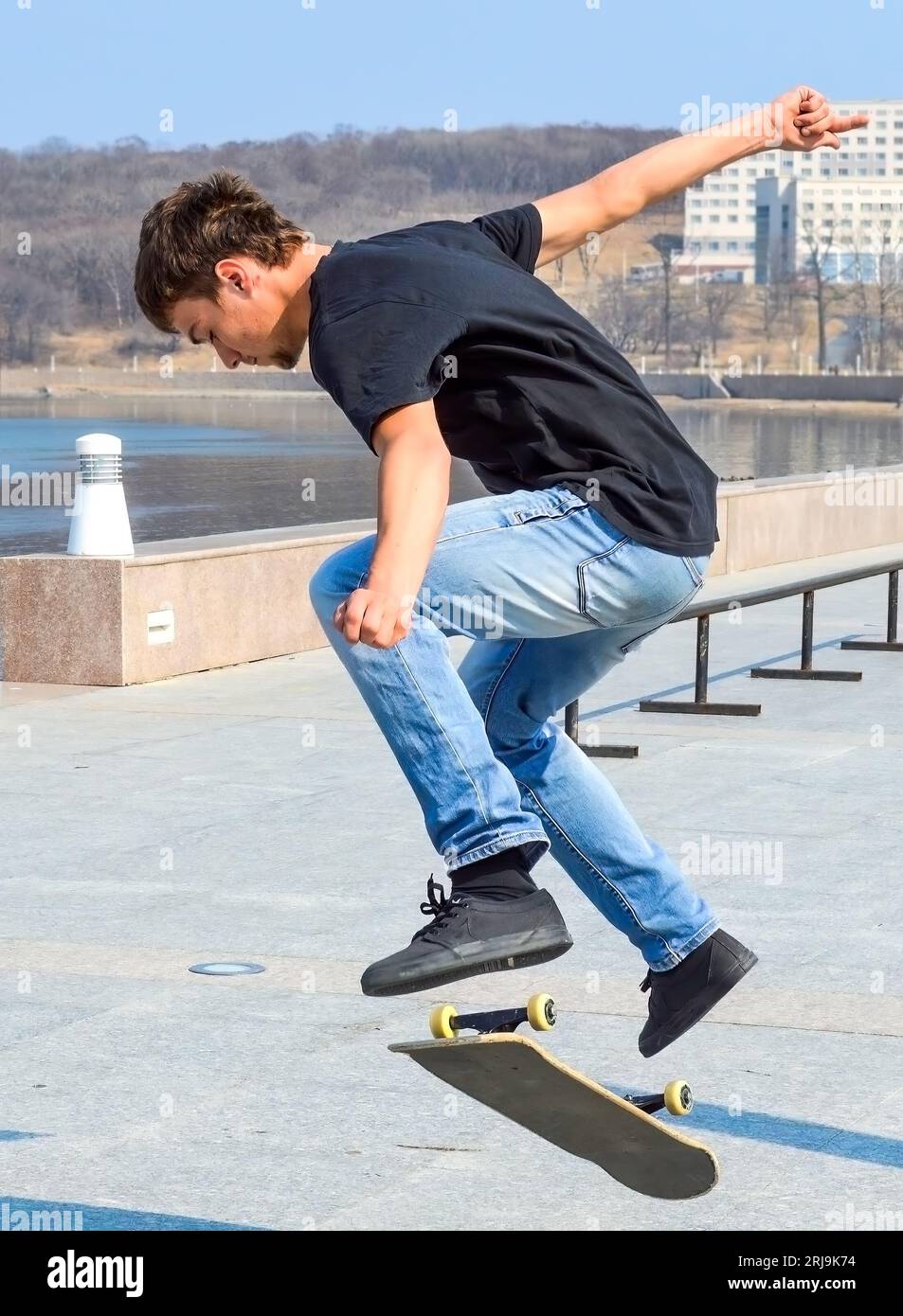Junger russischer Skater in schwarzem T-Shirt und blauer Jeans, der an einem frühen Frühlingstag auf dem Skateboard springt Stockfoto