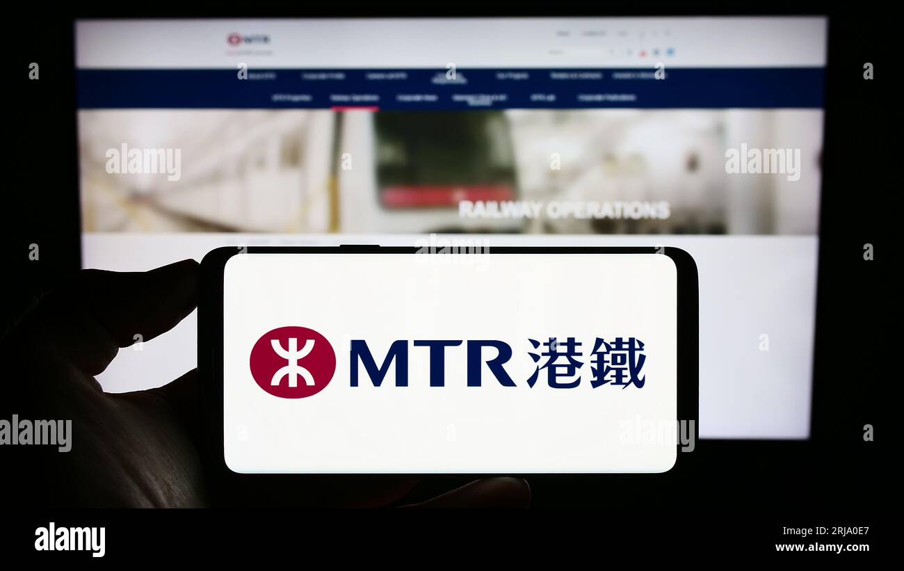 Person, die ein Mobiltelefon mit dem Logo der chinesischen Eisenbahngesellschaft MTR Corporation Limited auf dem Bildschirm vor der Webseite hält. Konzentrieren Sie sich auf das Display des Telefons. Stockfoto
