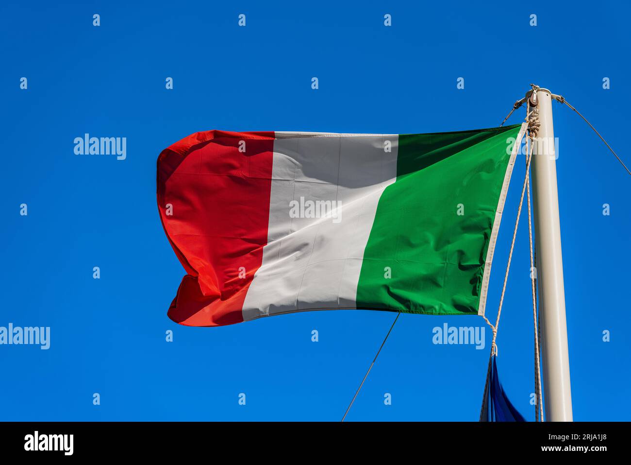 Nahaufnahme eines Mastes eines Segelbootes mit einer nationalen italienischen Flagge, die im Wind vor einem klaren blauen Himmel weht. Stockfoto