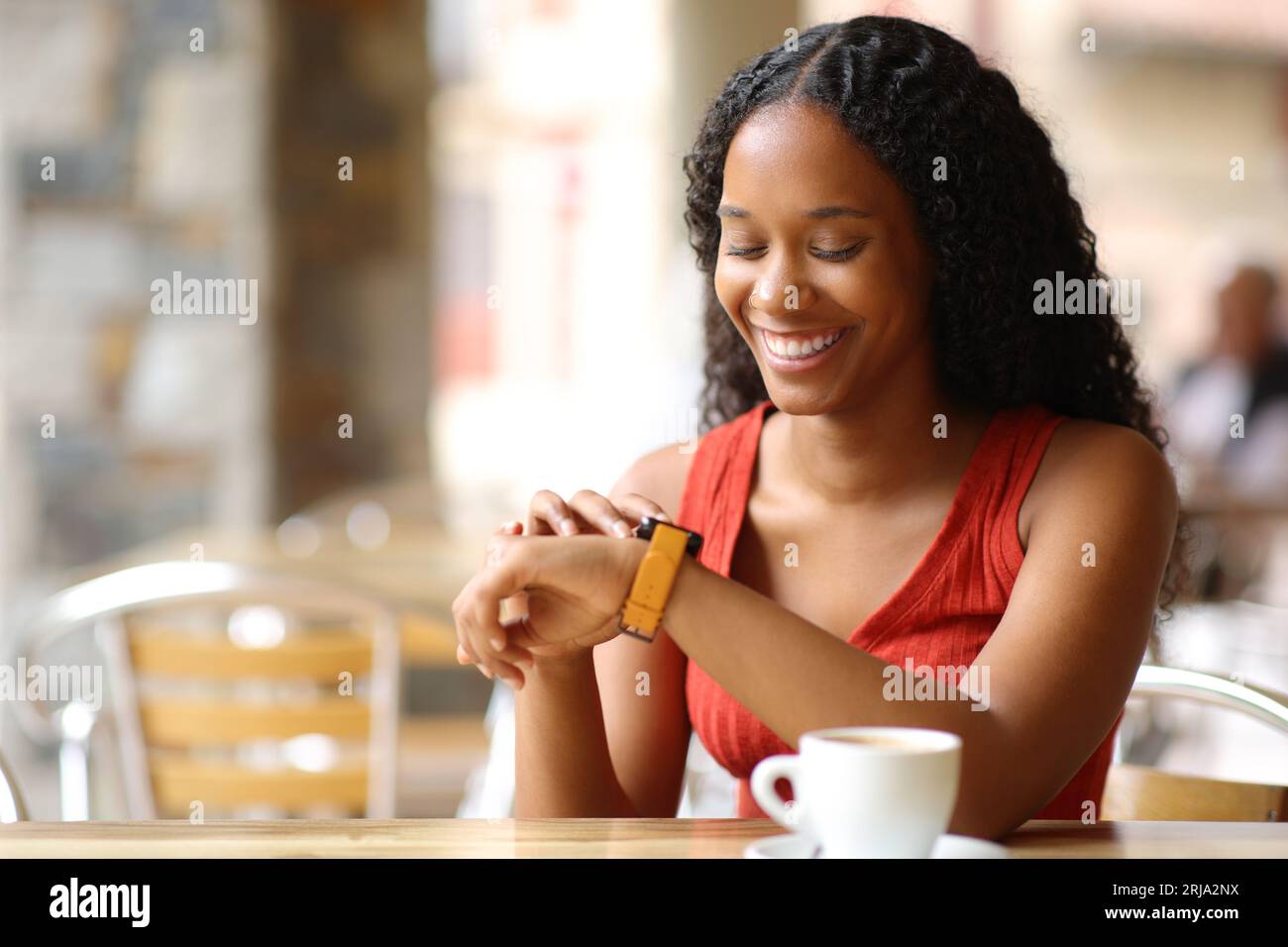 Glückliche schwarze Frau, die Smartwatch überprüft, sitzt in einer Café-Terrasse Stockfoto