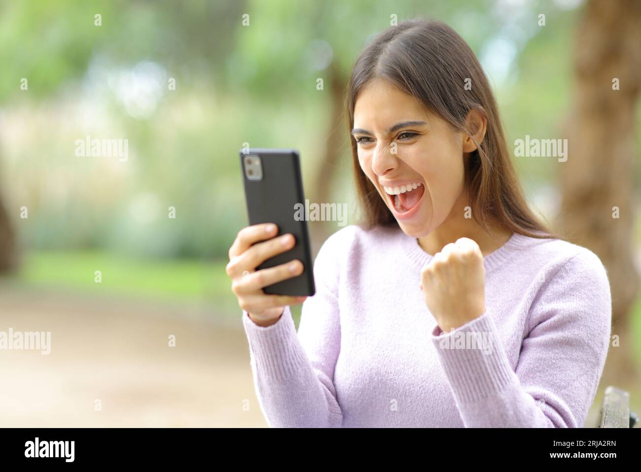 Aufgeregte Frau, die gute Nachrichten feiert und sich Telefoninhalte in einem Park ansieht Stockfoto