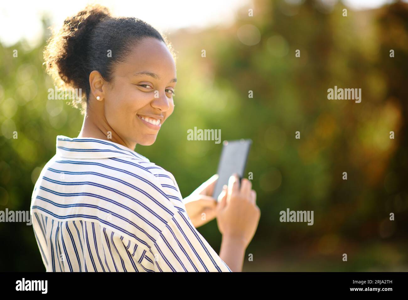 Glückliche schwarze Frau sieht dich an, wenn du dein Handy in einem Park hältst Stockfoto