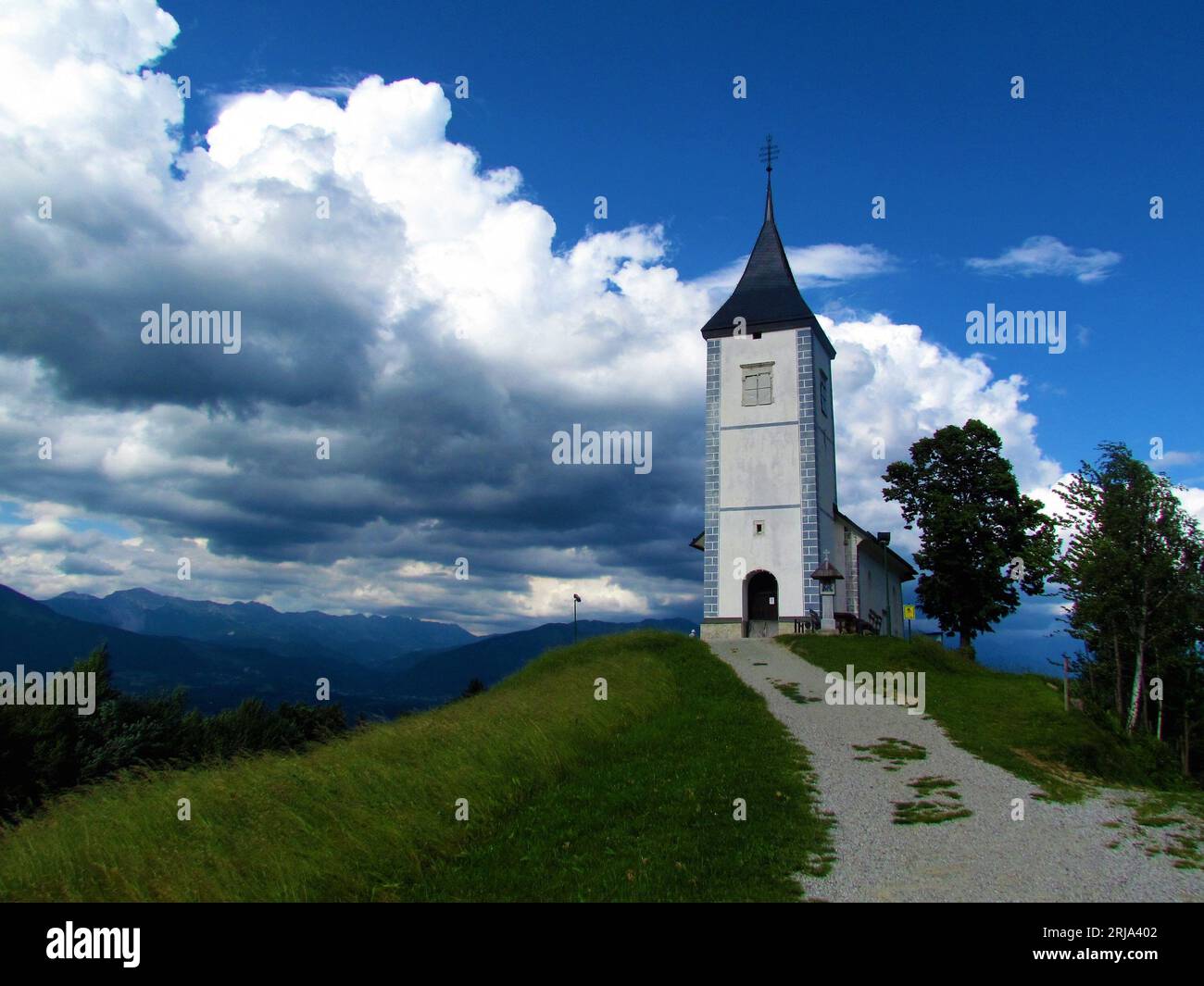 Kirche der Heiligen Primus und Felicius in Jamnik in Gorenjska, Slowenien mit dunklen Sturmwolken im Hintergrund und einer Schotterstraße, die in Richtung c führt Stockfoto