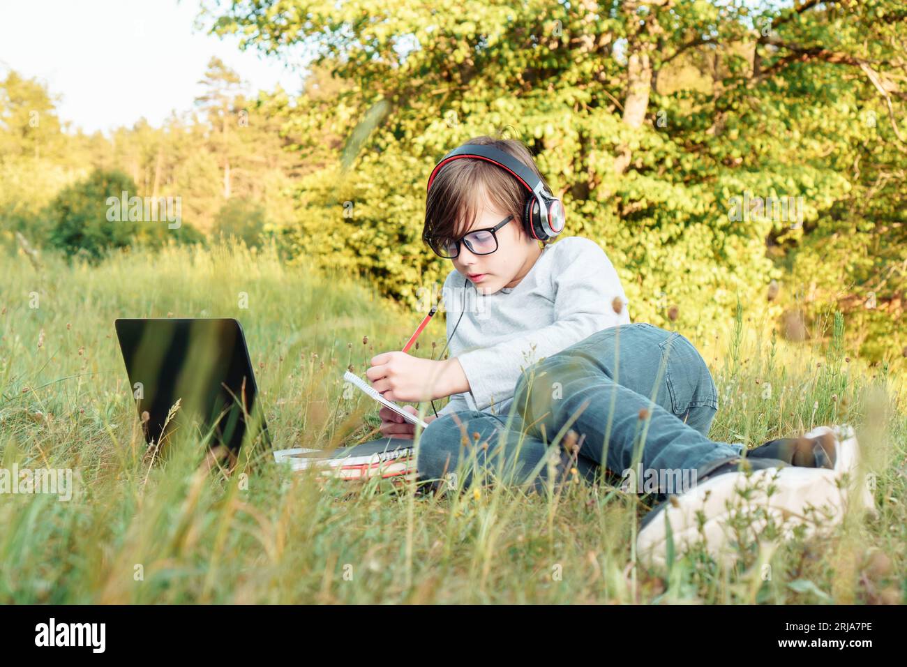 Konzentrierte Junior-Schülerin in Brille und Ohrhörern, die im Park vor dem Laptop auf grünem Gras liegt und Hausaufgaben im Notebook macht. Junge mit dunklem Haar Stockfoto