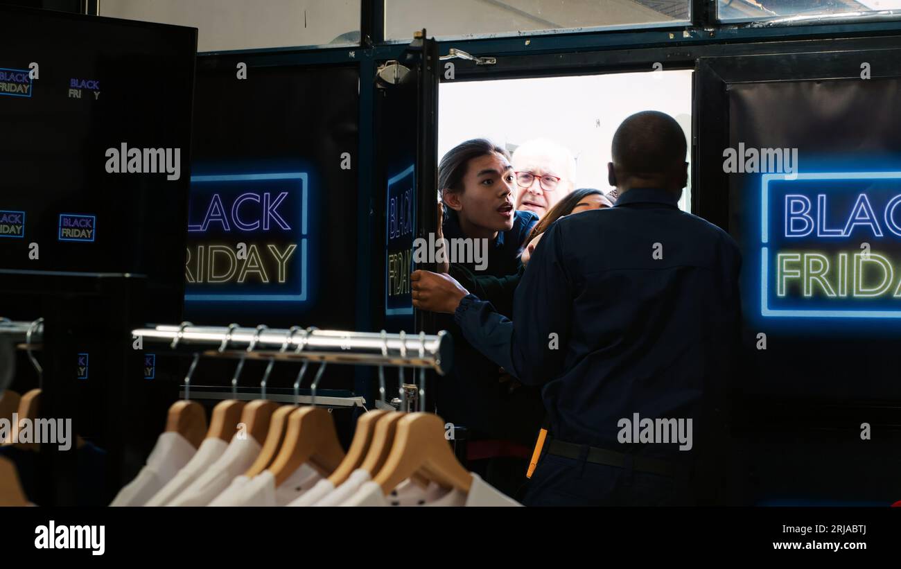 Kunden verlangen am schwarzen freitag einen Einkaufszugang und nutzen die frühen Öffnungszeiten, um billige Kleidung zu kaufen. Ängstliche, verrückte Gruppe von Leuten, die Tür drücken und mit dem Sicherheitsbeamten kämpfen. Handgeführte Aufnahme. Stockfoto