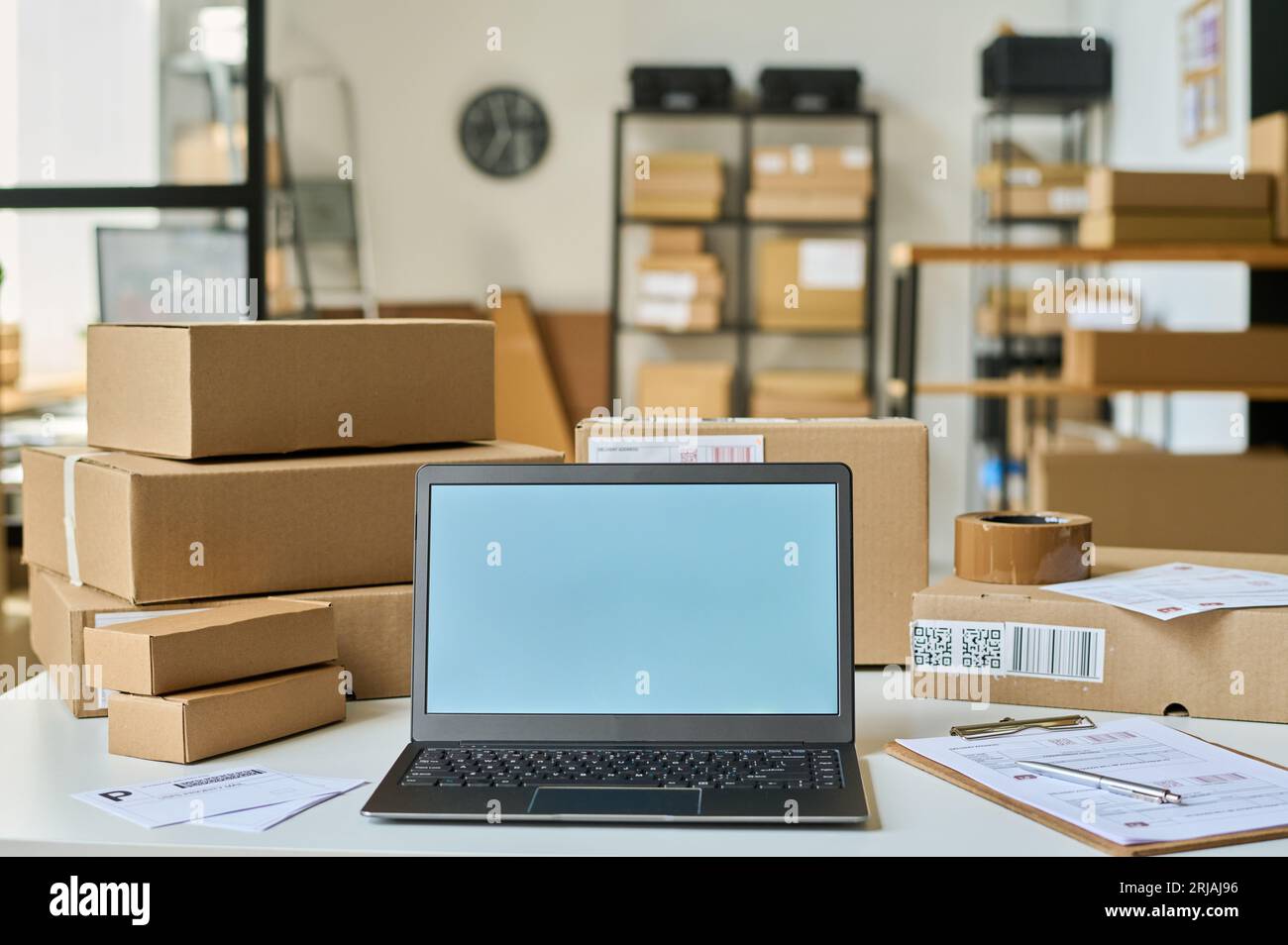 Laptop mit leerem Bildschirm, der zwischen Dokumenten steht, und verpackte Kartons, in denen Bestellungen von Online-Shops-Kunden im Lagerraum enthalten sind Stockfoto