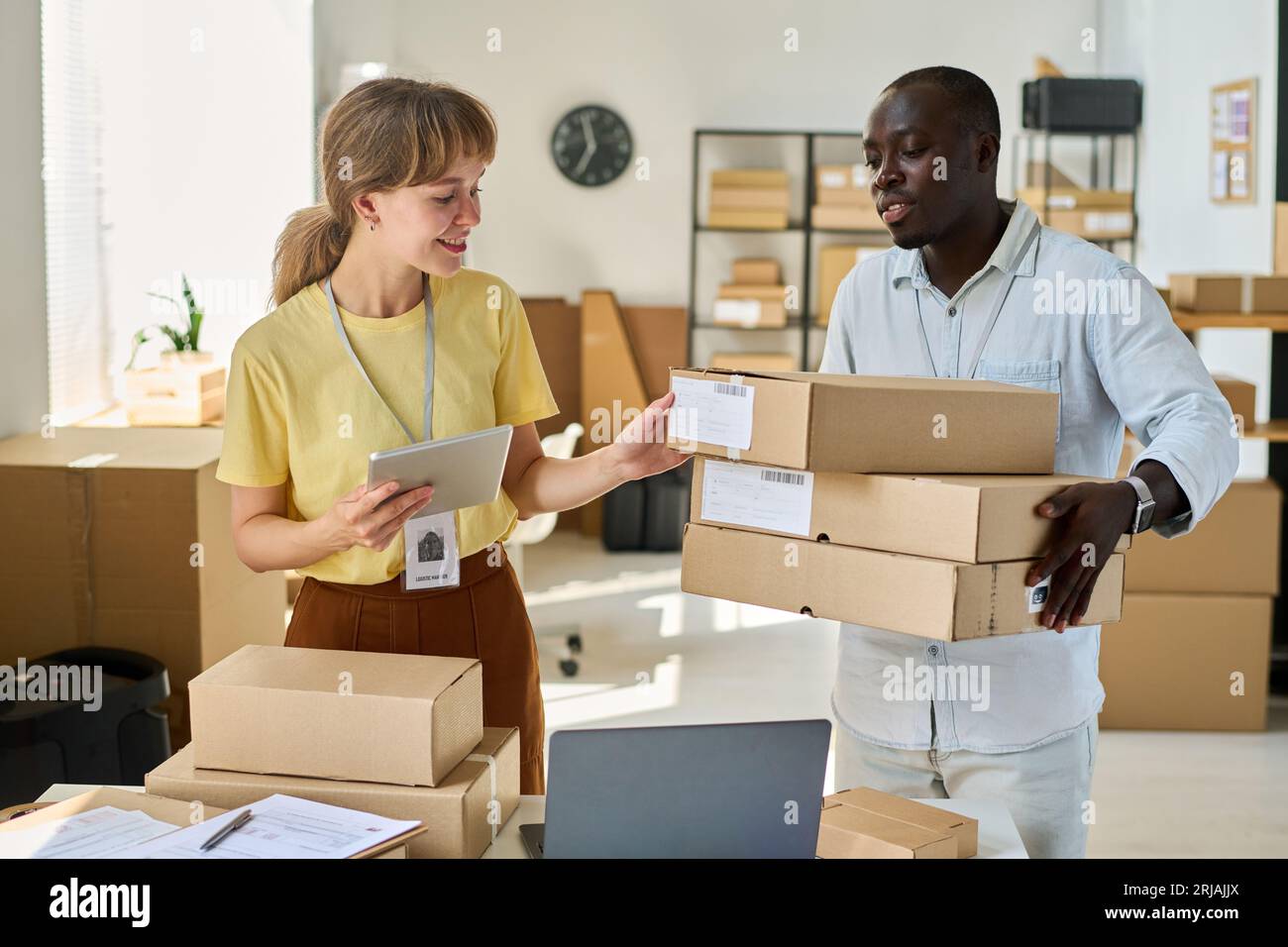 Glückliche junge Frau mit einem Tablet, das persönliche Daten von Kunden auf Kartons ihres afroamerikanischen männlichen Kollegen überprüft Stockfoto
