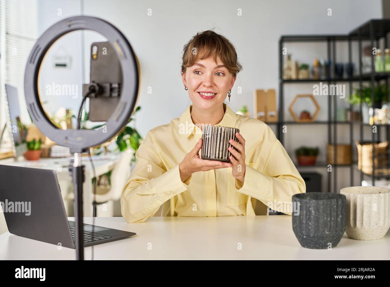 Glückliche junge Bloggerin sitzt am Schreibtisch vor dem Smartphone und macht Livestream von neuen Vasen oder Blumentöpfen aus dem Online-Shop Stockfoto